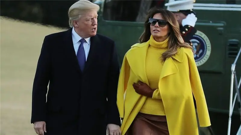 Дональд Трамп в черном пальто и Мелания Трамп в желтом пальто с коричневыми перчатками и солнцезащитными очками разговаривают и гуляют