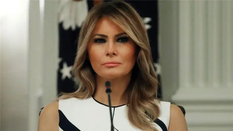 Мелания Трамп в бело-черном платье с нейтральным выражением лица перед микрофоном